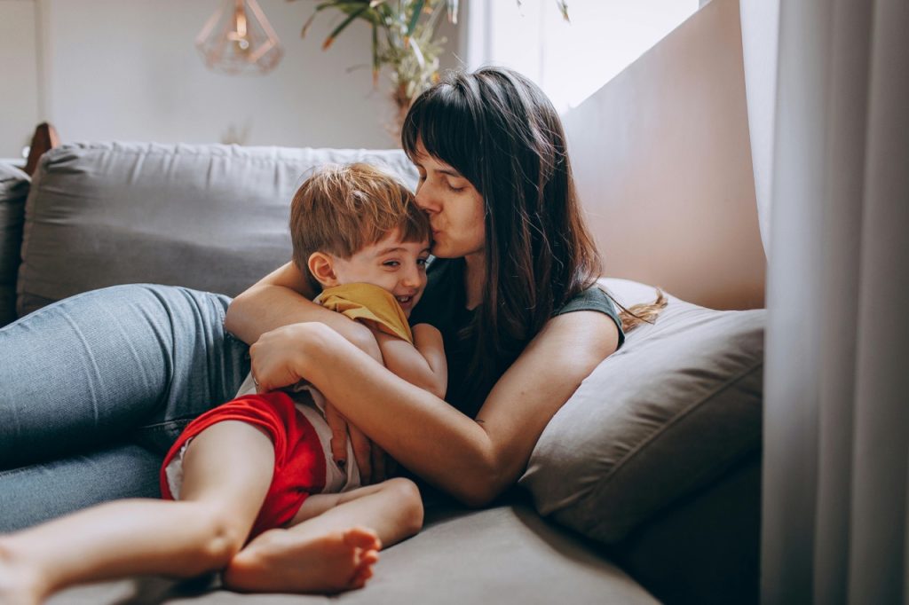 Mama i syn przytulają się na szarej kanapie, mama całuje syna w czoło